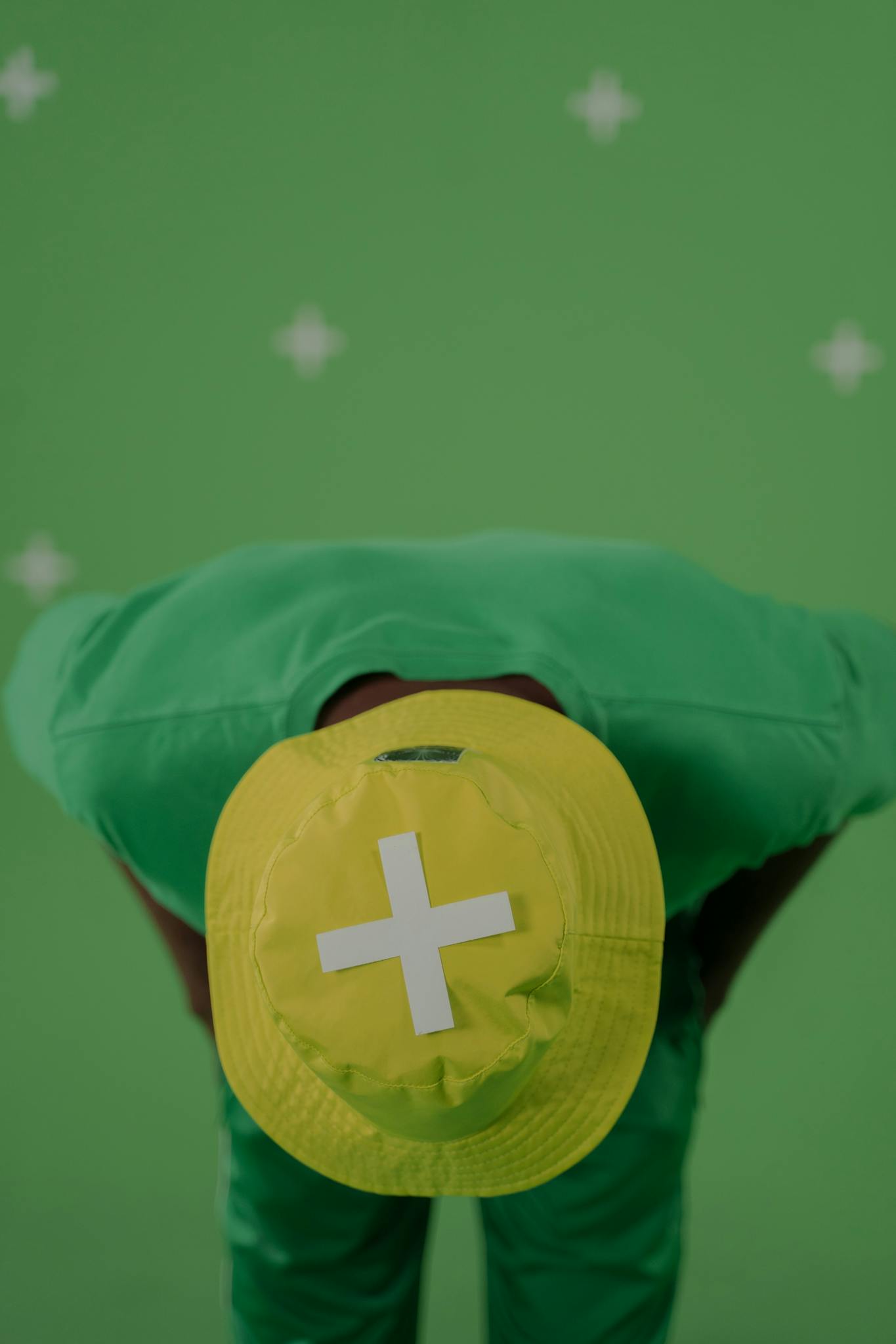 A person bending forward wearing a yellow bucket hat with cross against a green chroma key studio setup.