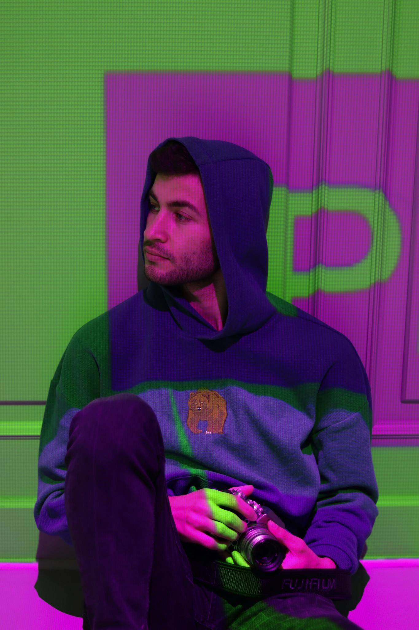 A stylish man in a hoodie jacket sitting indoors under colorful projector lights.